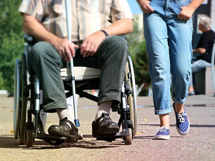 Arbeitsgemeinschaft Neues Leben Hennef e. V. | Bildausschnitt eines im Rollstuhl sitzenden Mannes und einer rechts daneben gehenden Frau in Jeans, sommerlich gekleidet
