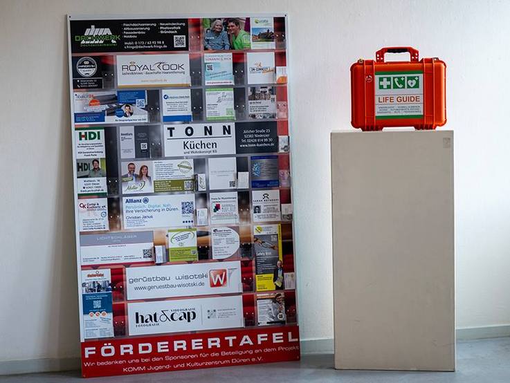 Förderertafel links vom weißen Sockel, auf dem der Lebensrettungs- und Notfallkoffer für das KOMM Kulturzentrum Düren steht