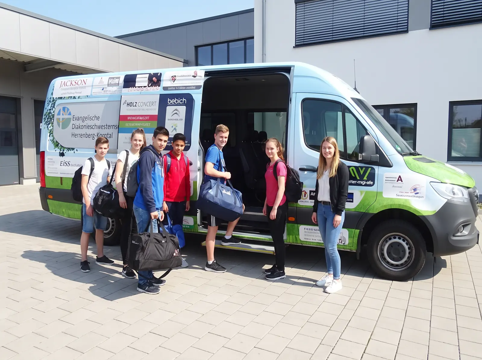 Jugendliche und eine Fachkraft beladen einen Kleinbus vor einer Einrichtung